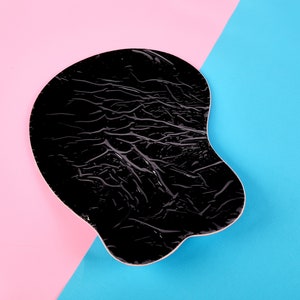 May include: A black ergonomic mouse pad with a textured surface. The pad is shaped to support the wrist and hand, and is set against a background of pink and blue.