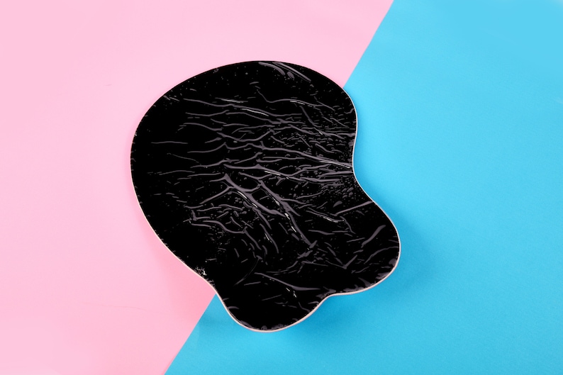 May include: A black ergonomic mouse pad with a textured surface. The pad is shaped to support the wrist and hand, and is set against a background of pink and blue.