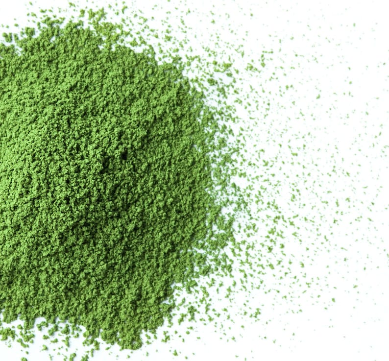 May include: A close-up of a pile of green matcha powder on a white surface. The powder is finely ground and has a light, airy texture.
