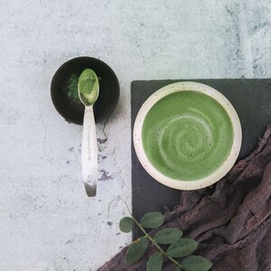 Puede incluir: Primer plano de un latte matcha verde en un taz&oacute;n blanco con un patr&oacute;n de remolino. Un taz&oacute;n peque&ntilde;o con polvo de matcha y una cuchara est&aacute; en el fondo.