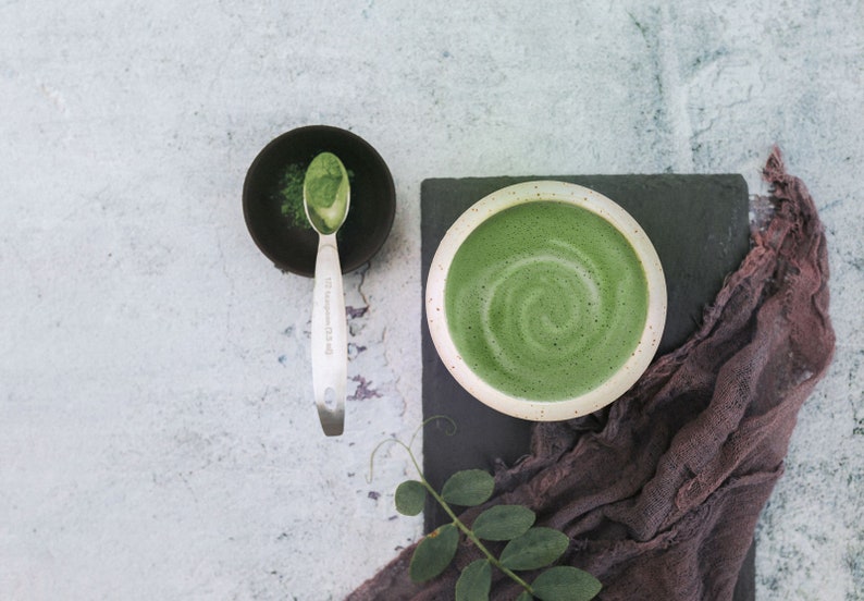 Puede incluir: Primer plano de un latte matcha verde en un taz&oacute;n blanco con un patr&oacute;n de remolino. Un taz&oacute;n peque&ntilde;o con polvo de matcha y una cuchara est&aacute; en el fondo.