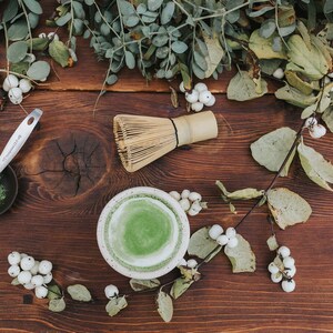 Puede incluir: Una mesa de madera con un cuenco de cer&aacute;mica blanca lleno de t&eacute; matcha verde, un batidor de bamb&uacute;, un peque&ntilde;o cuenco negro con polvo de matcha y una cuchara de plata. Bayas blancas y hojas verdes est&aacute;n esparcidas alrededor del cuenco.