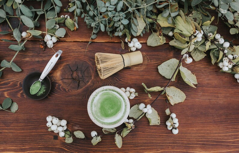 Puede incluir: Una mesa de madera con un cuenco de cer&aacute;mica blanca lleno de t&eacute; matcha verde, un batidor de bamb&uacute;, un peque&ntilde;o cuenco negro con polvo de matcha y una cuchara de plata. Bayas blancas y hojas verdes est&aacute;n esparcidas alrededor del cuenco.