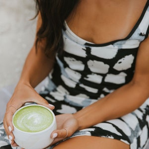 Puede incluir: Una persona sosteniendo una taza de cer&aacute;mica blanca llena de un latte matcha verde. La taza tiene un dise&ntilde;o de remolino verde claro en la parte superior del matcha. La persona lleva un vestido estampado en blanco y negro.