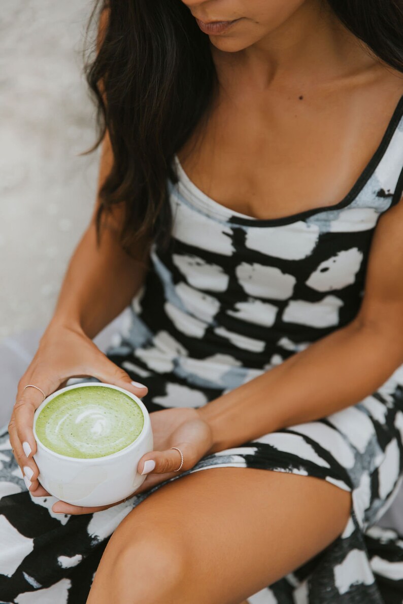 Puede incluir: Una persona sosteniendo una taza de cer&aacute;mica blanca llena de un latte matcha verde. La taza tiene un dise&ntilde;o de remolino verde claro en la parte superior del matcha. La persona lleva un vestido estampado en blanco y negro.