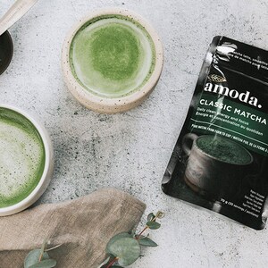 Puede incluir: Una bolsa de polvo de matcha verde de la marca "amoda." La bolsa tiene el texto "Classic Matcha" y "Energ&iacute;a limpia diaria y enfoque" en ella. Hay dos tazones de t&eacute; matcha verde sobre una superficie gris.