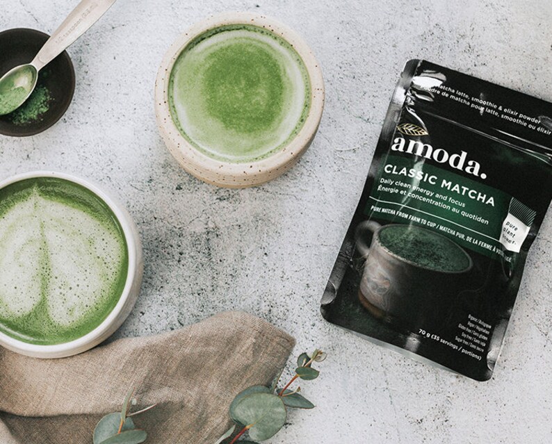 Puede incluir: Una bolsa de polvo de matcha verde de la marca "amoda." La bolsa tiene el texto "Classic Matcha" y "Energ&iacute;a limpia diaria y enfoque" en ella. Hay dos tazones de t&eacute; matcha verde sobre una superficie gris.