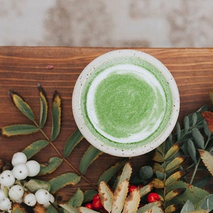 Puede incluir: Un bol de cer&aacute;mica blanca lleno de un batido verde vibrante, rodeado de ramitas de bayas verdes y blancas y hojas sobre una superficie de madera.