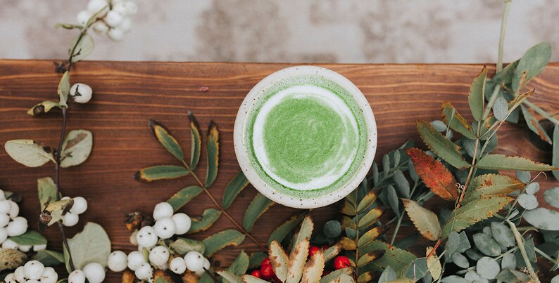 Puede incluir: Un bol de cer&aacute;mica blanca lleno de un batido verde vibrante, rodeado de ramitas de bayas verdes y blancas y hojas sobre una superficie de madera.