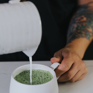 Puede incluir: Una taza de cer&aacute;mica blanca en la que se vierte un latte matcha verde. El latte tiene una parte superior espumosa.