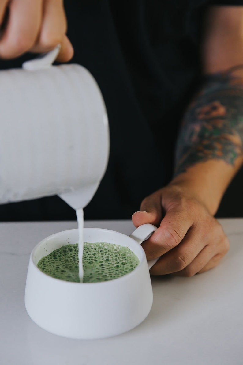 Puede incluir: Una taza de cer&aacute;mica blanca en la que se vierte un latte matcha verde. El latte tiene una parte superior espumosa.