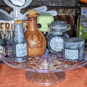 May include: A collection of five glass bottles with labels and lids, arranged on a clear glass pedestal. The bottles are decorated with a Halloween theme and contain various liquids. The labels read "Fog Juice", "Worms Wort", "Deadly Night Shade", "Frogs Breath", and "Jack Skellington".