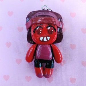 May include: A red and purple clay figurine of a cartoon character with a red headband, big eyes, and a toothy smile. The figurine is a keychain.