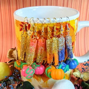 May include: A set of eight dangle earrings featuring a variety of colors and glitter. Each earring has a rectangular resin charm with a pumpkin charm attached to the bottom. The charms are in shades of orange, pink, blue, and purple.
