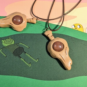 May include: Two tan-colored pendant necklaces with a brown stone center, one with a cord. The pendants are shaped like a stylized head. The background features cartoon characters and a green and pink color scheme.