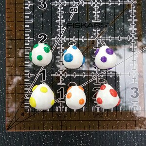 以下が含まれることがあります： 異なる色の斑点が付いた白い卵型のチャームが6つ。チャームはポリマークレイ製で、吊り下げ用の銀色のループが付いています。斑点の色は緑、青、紫、黄、オレンジ、赤です。