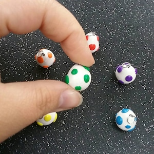 以下が含まれることがあります： 異なる色の水玉模様がついた、小さな白い卵型のチャームが6つ。チャームには、吊り下げ用の銀色のジャンプリングが付いています。