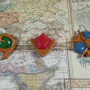 May include: Three ceramic pins with a map background. The pins are shaped like a green swirl, a red star, and a blue and brown spider.