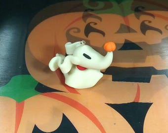 May include: A white clay figurine of a ghost with a small orange pumpkin on its head. The ghost has a black eye and a red mouth. The figurine is sitting on a brown and orange surface.