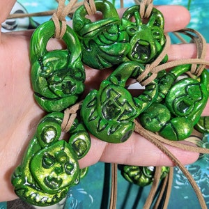 May include: A set of six green, hand-painted, ceramic pendants with a metallic sheen. Each pendant features a different design, including a skull, a heart, and a flower. The pendants are strung on brown leather cords.