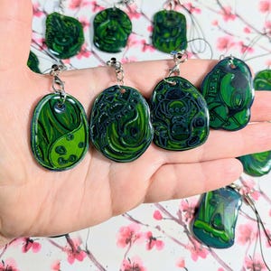 Puede incluir: Una colección de colgantes verdes y azul oscuro con llaveros plateados. Los colgantes tienen varios diseños, incluyendo patrones abstractos y un símbolo de yin-yang. Los colgantes se sostienen en una mano, con un fondo floral.