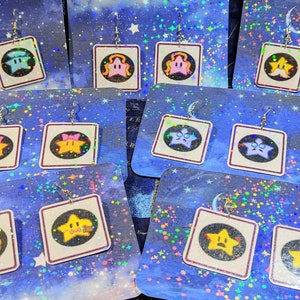 May include: A set of 10 pairs of star-shaped earrings with different colored faces. The earrings are on cards with a glittery blue background and white stars.