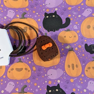 May include: A brown chocolate chip cookie with an orange bow tie charm on a brown cord necklace. The necklace is on a purple background with a Halloween theme featuring pumpkins, ghosts, and black cats.