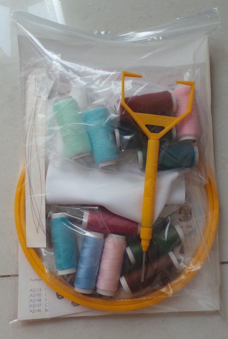 May include: A clear plastic bag containing a variety of embroidery floss spools in different colors, a yellow embroidery hoop, a white fabric square, and a yellow plastic needle threader. The bag also includes a piece of paper with the text "Further A2113 1 A2150 / A2148 S A2147 C A2146 Br".