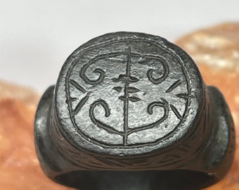 Puede incluir: Un anillo de metal negro con un diseño circular que presenta un símbolo estilizado. El anillo está grabado con detalles intrincados y tiene una superficie texturizada. El texto "OTHER TIME RINGS" y "ANCIENT ARTIFACTS AND RESOURCES" es visible sobre el anillo.