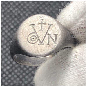 Pu&ograve; includere: Un anello a sigillo d'argento con una faccia rotonda che presenta una croce e le lettere "V" e "N".