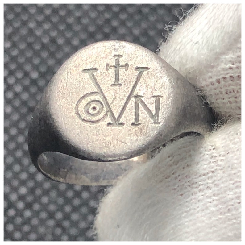 Pu&ograve; includere: Un anello a sigillo d'argento con una faccia rotonda che presenta una croce e le lettere "V" e "N".