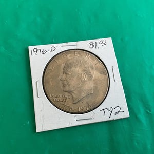 May include: A 1976-D Eisenhower dollar coin in a protective holder. The coin features a profile of Dwight D. Eisenhower and the text "IN GOD WE TRUST" and "1776-1976". The holder has handwritten text "1976-D" and "TY2".