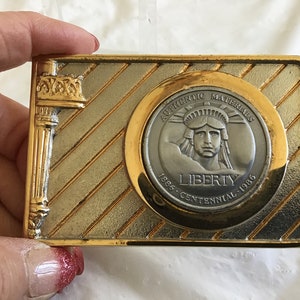 May include: A gold-toned belt buckle with a silver-toned medallion featuring a depiction of the Statue of Liberty. The medallion has the text "Authentic Materials 1886 - Centennial - 1986" and "Liberty" around the image.