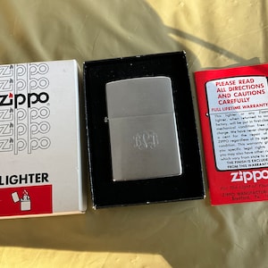May include: A silver Zippo lighter with a monogrammed design. The lighter is in its original box with a red and white Zippo logo. The box also includes a warranty card with instructions for use.