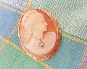 May include: A gold-toned cameo brooch with a white cameo of a woman's profile. The cameo is set in a gold-toned oval frame with a diamond accent.