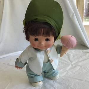 May include: A vintage doll dressed in a white jacket, light blue pants, and a green hat. The doll is holding a pink ice cream cone. The doll has brown hair and a smiling face. The doll's shoes are white.