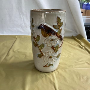 May include: A ceramic vase with a white background and brown and green floral design. A brown bird with a yellow breast is perched on a branch with white flowers.