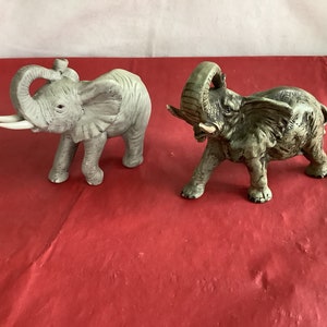 May include: Two ceramic elephant figurines. One is gray and the other is a darker gray with brown accents. Both elephants have their trunks raised.