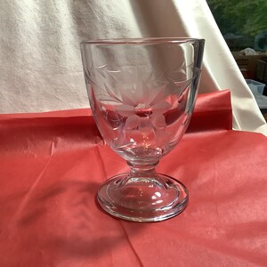 May include: A clear glass goblet with a floral etched design. The goblet has a wide bowl and a tall, slender stem with a round base.