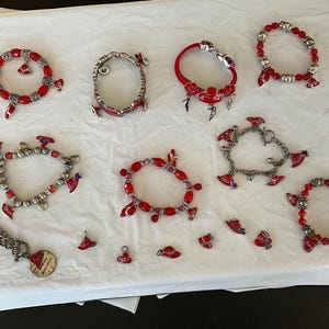 May include: A collection of bracelets and charms with red and silver accents. The bracelets feature red beads and decorative charms. Some bracelets are silver with red and purple charms. A silver chain necklace with a round charm is also present.