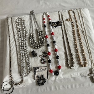 May include: A collection of silver and white beaded necklaces, including a black and white patterned necklace with red beads, and a silver cross pendant necklace. There are also earrings with black and white beads and a tag that says "Dearly Diamonds".
