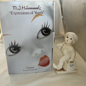 May include: A white porcelain figurine of a young boy carrying a satchel, next to a box with the text "M.J. Hummel Expressions of Youth". The figurine is on a small rectangular base. The box features a close-up of a doll's face.
