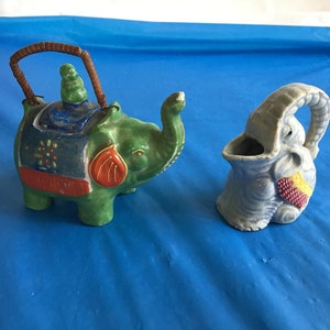 May include: Two ceramic elephant figurines. One is green with red, blue, and gold accents and has a brown handle. The other is blue with red and yellow accents.