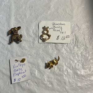 May include: A collection of vintage gold-toned brooches and pins. Includes a dog, a bunny with a pearl, an elephant, and a mouse. The bunny brooch is labeled "Rhinestone Bunny Pearl Tail". The elephant pin is labeled "Vintage Baby Elephant".
