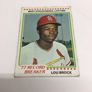 May include: A vintage baseball card featuring Lou Brock, a baseball player for the St. Louis Cardinals. The card has a red, white, and blue color scheme and features the text "'77 Record Breaker" and "Most Stolen Bases, Lifetime".