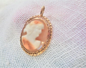 May include: A gold-toned pendant featuring a cameo of a woman's profile in a shell-like setting. The cameo is surrounded by a delicate, intricate gold filigree design.