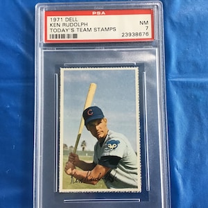 May include: A 1971 Dell Today's Team Stamps baseball card featuring Ken Rudolph, a Chicago Cubs player. The card is graded NM 7 by PSA and has the number 23938676.