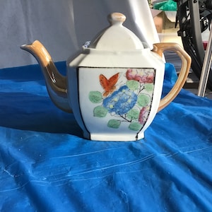 May include: A white teapot with a brown handle and spout. The teapot has a floral design with a butterfly in the center. The flowers are blue, pink, and green.