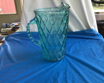 May include: A light blue glass pitcher with a diamond pattern. The pitcher has a handle and a spout.