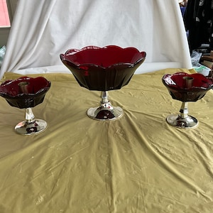 May include: A set of three ruby red glass candle holders and a centerpiece bowl. The candle holders have a scalloped edge and sit on a silver base. The centerpiece bowl has a similar design and is larger.